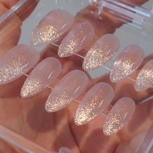 May include: A set of almond-shaped, press-on nails. The nails are a pale pink with a glitter gradient, transitioning from a sparkly gold at the tips to a translucent base. They are arranged in a clear plastic case.