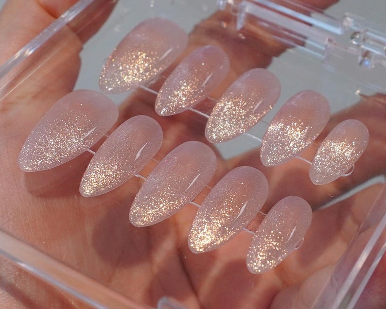 May include: A set of almond-shaped, press-on nails. The nails are a pale pink with a glitter gradient, transitioning from a sparkly gold at the tips to a translucent base. They are arranged in a clear plastic case.