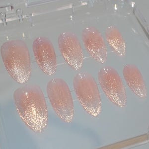 May include: A set of ten almond-shaped, press-on nails. The nails have a gradient design, transitioning from a light pink base to a glittery gold tip. They are arranged in a clear plastic case.