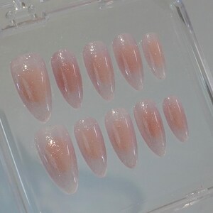 May include: A set of ten almond-shaped false nails. Each nail is a gradient of pale pink to clear, with a glittery shimmer. The nails are arranged in a clear plastic case.