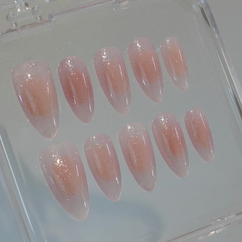 May include: A set of ten almond-shaped false nails. Each nail is a gradient of pale pink to clear, with a glittery shimmer. The nails are arranged in a clear plastic case.