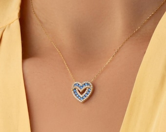 Op de afbeelding: Een gouden ketting met een hartvormige hanger. De hanger is ontworpen met een buitenrand van heldere witte stenen en een binnenste hartvorm gevuld met felblauwe stenen, wat zorgt voor een gelaagd en gedetailleerd uiterlijk.