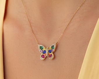 Op de afbeelding: Een gouden ketting met een vlinderhanger. De vlindervleugels zijn versierd met kleine, druppelvormige edelstenen in rood, blauw, groen en geel, met een centrale rij heldere stenen en een rand van kleine pavé-kristallen.