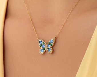 Op de afbeelding: Een gouden ketting met een vlinderhanger. De vlindervleugels zijn versierd met kleine, ronde, lichtblauwe en groene stenen, geaccentueerd door kleine heldere kristallen langs de randen van de vleugels en het lichaam van de vlinder.