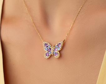 Op de afbeelding: Een gouden ketting met een vlindervormige hanger. De hanger is ingelegd met kleine, paarse, druppelvormige stenen en kleine, heldere, sprankelende accentstenen langs de randen en in het midden van de vlindervleugels.