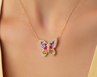 Op de afbeelding: Een gouden ketting met een vlindervormige hanger. De hanger is aan de randen bezet met kleine heldere steentjes en bevat meerdere gekleurde, gefacetteerde stenen in blauwe, roze, rode en groene tinten in de vleugels.