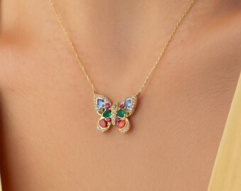 Op de afbeelding: Een gouden ketting met een vlinderhanger. De vleugels van de vlinder zijn versierd met kleine, kleurrijke edelstenen in blauwe, groene en rode tinten, en de randen van de vleugels zijn bezet met piepkleine, heldere, fonkelende witte steentjes.