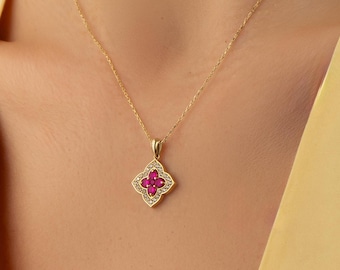 Op de afbeelding: Een gouden ketting met een hanger in de vorm van een bloem met vier bloemblaadjes. De hanger is in het midden bezet met vier kleine, felroze stenen, omringd door een rand van kleine, heldere, fonkelende steentjes in een gouden frame.