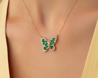 Op de afbeelding: Een gouden ketting met een vlindervormige hanger. De hanger is versierd met meerdere peervormige groene stenen en kleine heldere steentjes langs de randen, wat een gedetailleerd vleugelpatroon vormt aan een fijne gouden metalen schakelketting.