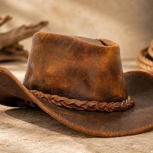 May include: A brown leather cowboy hat with a distressed finish and a braided leather band around the base of the crown. The hat features a wide, curved brim and a structured, tall crown, showcasing natural variations in the leather texture.