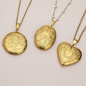 Könnte beinhalten: Drei goldfarbene Medaillon-Halsketten. Eine ist rund mit floralem Design, eine andere ist oval mit floralem Design und die dritte ist herzförmig mit floralem Design. Jedes Medaillon hängt an einer zarten Kette.