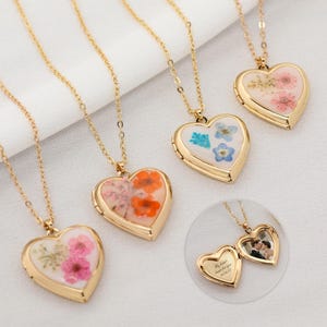 May include: Gold-toned heart-shaped lockets with delicate floral arrangements inside, displayed on gold chains. The lockets contain pink, orange, and blue dried flowers. A small, open locket shows a photo and text.