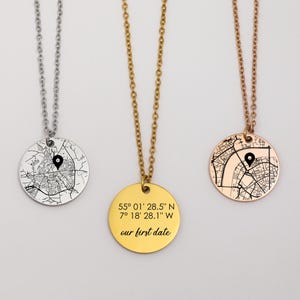 May include: Three necklaces with round pendants. One is silver, one is gold, and one is rose gold. Each pendant features a map design with a black location marker. The gold pendant has the text "55° 01' 28.5" N 7° 18' 28.1" W our first date."