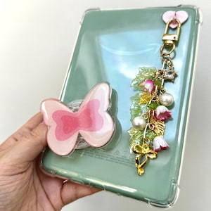 May include: A clear tablet case with a pink butterfly phone grip and a gold charm. The charm features a pink heart, a star, faux pearls, and pink and white flower accents. The tablet is light green.
