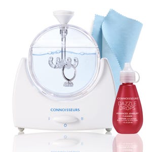 May include: A white jewelry cleaner with a clear, round cleaning chamber holding a ring. A red bottle of "Dazzle Drops" jewelry cleaner and a blue cleaning cloth are also included. The cleaner has the brand name "Connoisseurs" printed on it.