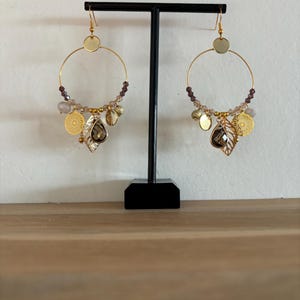 Op de afbeelding: Gouden oorringen met bungelende bedels. Elke oorbel heeft een grote gouden ring met kleine paarse kralen, gouden bladbedels en een druppelvormig kristal. De oorbellen worden op een zwarte standaard gepresenteerd.