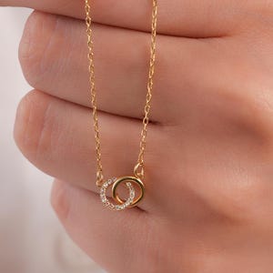 Pode incluir: Um colar de tom dourado com uma corrente delicada e um pendente composto por dois anéis circulares entrelaçados. Um anel é de metal liso de tom dourado, enquanto o outro está adornado com pequenas pedras claras e brilhantes no bordo.