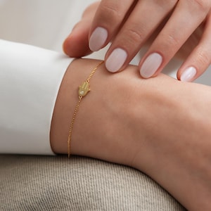 May include: Gold-colored bracelet featuring a Hamsa hand charm with a small, clear gemstone. The bracelet is delicate, with a fine chain. The charm is centered on the wrist, and the bracelet is worn against a white shirt.