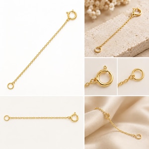 May include: A gold-toned jewelry extender chain featuring a circular spring ring clasp at one end and a small jump ring at the other. The delicate metal chain is shown in multiple views, highlighting the polished finish and the functional clasp mechanism.