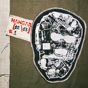 May include: A black and white skull-shaped patch with a black border and white stitching. The skull is filled with a collage of images. A small rectangular tag with the text "HANGAR [12|13] #1" is attached.