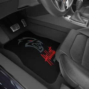 May include: Black car floor mat featuring the Hellcat logo in gray and red. The logo depicts a stylized demon head with red eyes. The mat is placed in the driver's side footwell of a car, near the pedals.