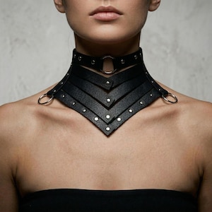 May include: A black leather choker necklace with a geometric design. The choker features multiple layers, silver-tone rivets, and a large silver ring. The necklace is designed to sit high on the neck and has a bold, edgy aesthetic.