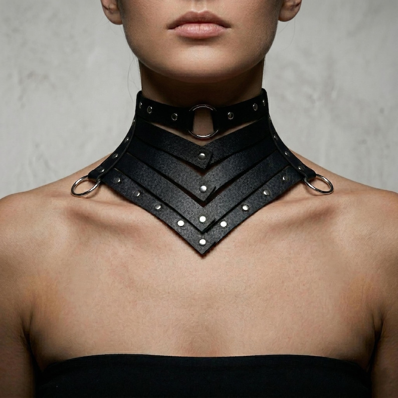 May include: A black leather choker necklace with a geometric design. The choker features multiple layers, silver-tone rivets, and a large silver ring. The necklace is designed to sit high on the neck and has a bold, edgy aesthetic.