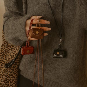 Könnte beinhalten: Kleine Lederetuis in Rot, Braun und Schwarz, jeweils mit Knopfverschluss. Die Etuis sind an dünnen Kordeln befestigt, was darauf hindeutet, dass sie als Halsketten getragen oder an anderen Gegenständen befestigt werden können. Im Hintergrund ist eine Tasche mit Leopardenmuster zu sehen.