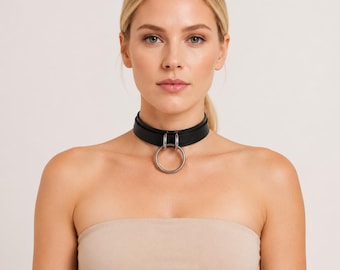 May include: A black leather choker necklace featuring a smooth, wide band that sits snugly around the neck. The center of the choker is adorned with a large, circular silver-toned metal ring attached to the band by two smaller silver-toned loops.