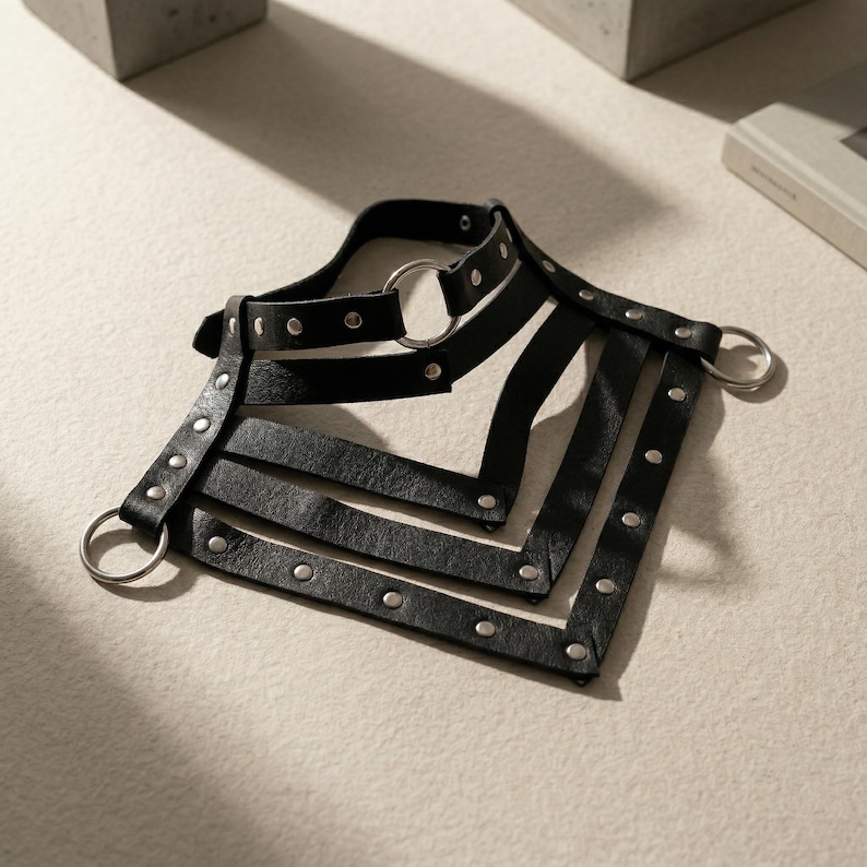 May include: A black leather harness with silver-toned metal rings and rivets. The harness features a collar, shoulder straps, and chest straps, all connected with metal rings. The design is symmetrical and angular, with a minimalist aesthetic.