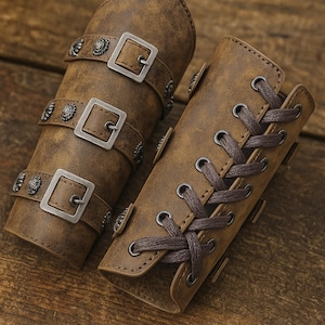 Puede incluir: Brazaletes de cuero marrón con hebillas plateadas y tachuelas decorativas. Un brazalete tiene tres hebillas, el otro un diseño con cordones. Los brazaletes probablemente son parte de un disfraz o un conjunto de accesorios.