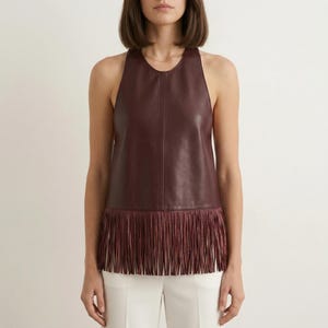 Può includere: Un top senza maniche in pelle bordeaux con orlo a frange. Il top ha una scollatura rotonda e una silhouette aderente. La frangia è dello stesso colore del top. La modella indossa pantaloni bianchi.