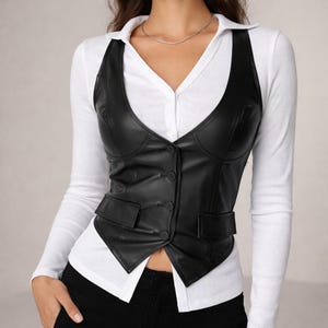 May include: A black faux leather vest with a V-neckline and a button-up front. The vest has two small pockets and is worn over a white collared shirt. The vest is a stylish addition to any wardrobe.