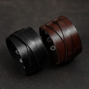 Puede incluir: Dos pulseras de cuero para la muñeca. Una es negra, la otra marrón. Ambas tienen un diseño tejido y cierres de botón a presión plateados. Las pulseras son un accesorio audaz, adecuado para varios estilos.