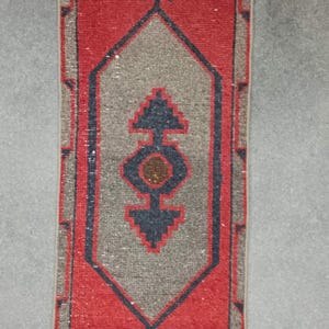 May include: Rectangular, hand-woven rug with a red border and a central design in grey and navy blue. The rug features a geometric pattern with a central medallion and decorative corner elements. The rug is approximately 120 cm long.