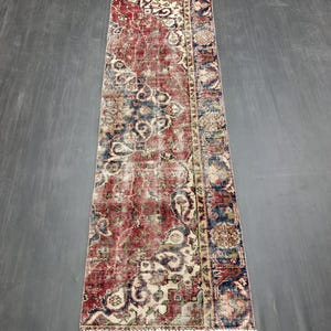 Puede incluir: Una alfombra rectangular de estilo vintage con aspecto desgastado. La alfombra presenta un patrón complejo en tonos de rojo, crema y azul marino. El diseño incluye elementos florales y geométricos, con un borde con flecos en un extremo. La alfombra está colocada sobre una superficie gris oscuro.