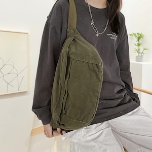 May include: An olive green canvas sling bag with a single shoulder strap. The bag has a rectangular shape with a zippered pocket on the front. The bag is worn across the body. The person is wearing a dark grey long-sleeved top and light grey trousers.