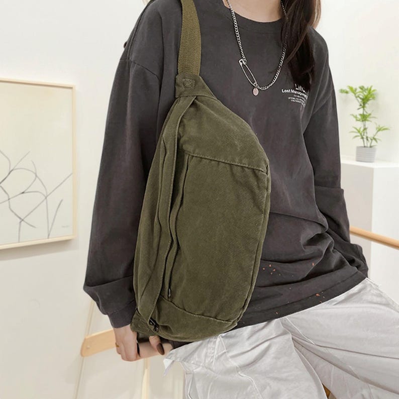 May include: An olive green canvas sling bag with a single shoulder strap. The bag has a rectangular shape with a zippered pocket on the front. The bag is worn across the body. The person is wearing a dark grey long-sleeved top and light grey trousers.