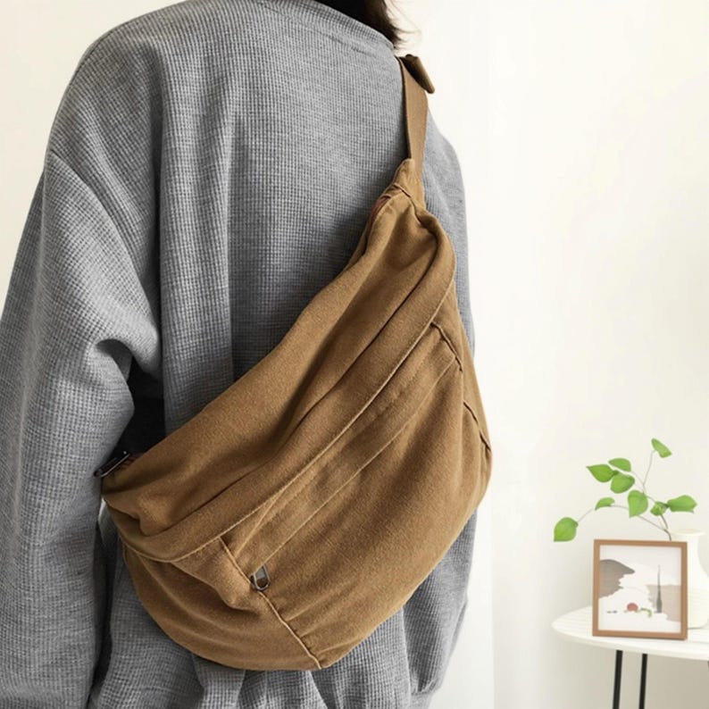 May include: A brown canvas crossbody bag with a single shoulder strap. The bag has a zippered pocket on the front and a curved shape. The bag is worn over the shoulder.