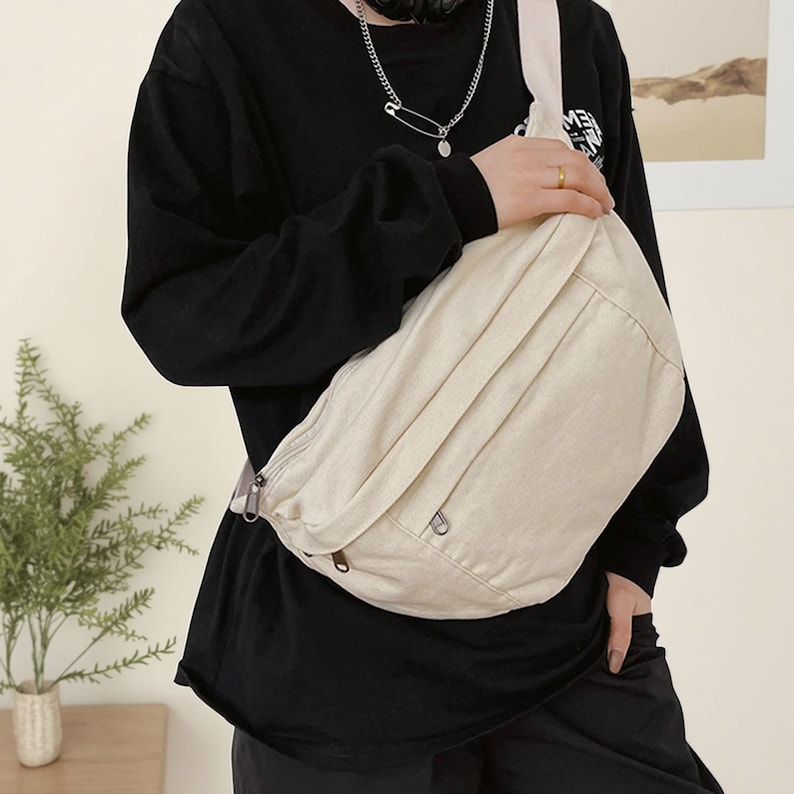May include: A cream-coloured crossbody bag with a zip-up main compartment and an adjustable strap. The bag is made of a canvas-like material and has a minimalist design. The bag is worn over the shoulder.