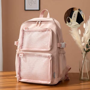 May include: A pale pink backpack featuring two front mesh pockets and a top handle. The backpack has adjustable straps and a buckle fastening. The word "Jack" is printed on the shoulder strap. The backpack is on a wooden surface.