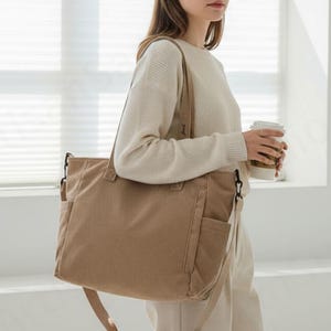 Peut inclure: Un sac fourre-tout en velours côtelé beige avec une bandoulière et plusieurs poches extérieures. Le sac est de forme rectangulaire et possède une poignée supérieure. Le sac est tenu par une personne.