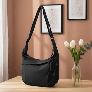 May include: Black shoulder bag with a long adjustable strap. The bag has a flap closure and a small zipper pocket. The word "Jack" is printed on the strap. The bag is sitting on a wooden surface.