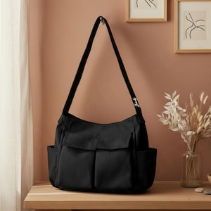 May include: A black shoulder bag with two front pockets and a long strap. The word "Jack" is printed in white on the strap. The bag is hanging on a hook against a peach-colored wall.