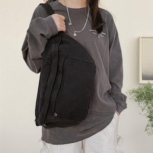 May include: A black canvas crossbody bag with a large front pocket and adjustable strap. The bag is worn over the shoulder. The person is wearing a grey long-sleeve shirt and white trousers. A silver necklace is visible.