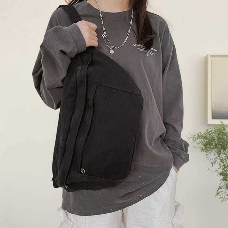 May include: A black canvas crossbody bag with a large front pocket and adjustable strap. The bag is worn over the shoulder. The person is wearing a grey long-sleeve shirt and white trousers. A silver necklace is visible.