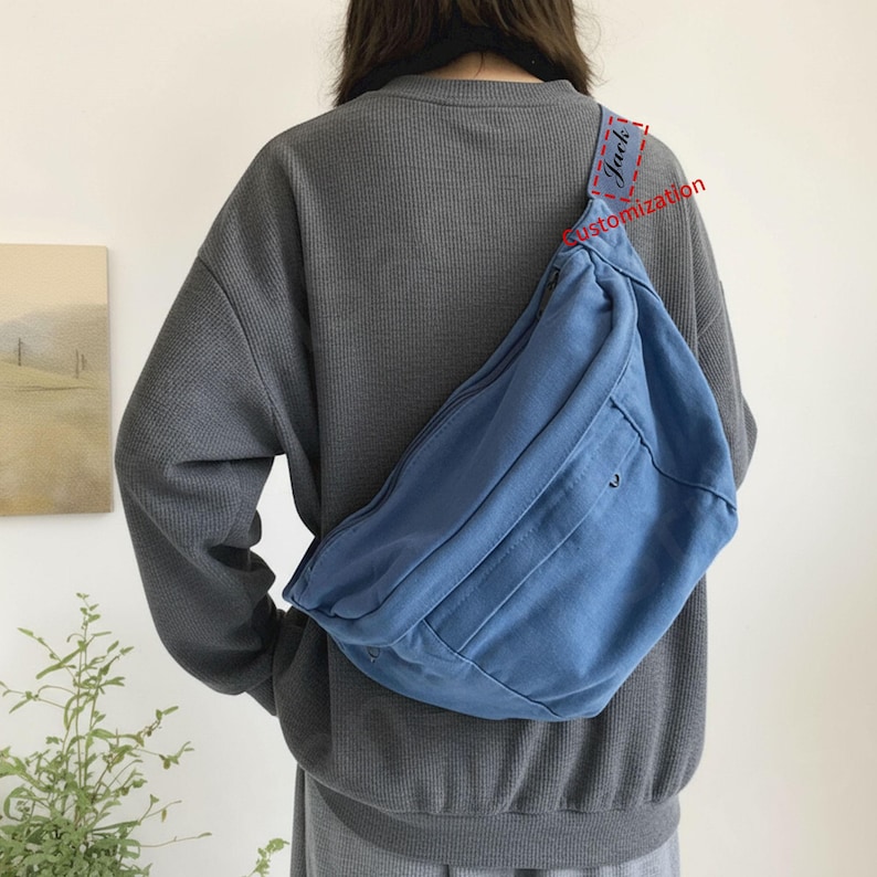 May include: A light blue canvas crossbody bag with a large main compartment and a smaller front pocket. The bag is worn over the shoulder. The word "Jack" is visible on the strap. The bag is a light blue colour.