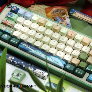 May include: A custom mechanical keyboard with a green frame and keycaps featuring various illustrations. The keycaps have a mix of colors, including white, green, and blue, with images of landscapes and characters. The keyboard has a wrist rest with a blue and white design. The text "KEYBOARD//KRAFT" is visible.