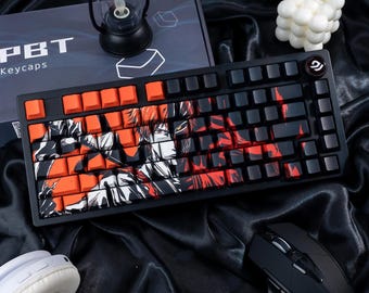 May include: Black and orange mechanical keyboard with a graphic design. The keyboard has orange keycaps on the top row and a black and red design on the main keys. A black mouse and white headphones are also in the image. The box reads "PBT Keycaps".