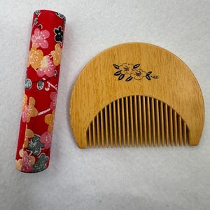 May include: A semi-circular wooden comb with a black floral line drawing and a cylindrical case covered in red fabric featuring a multi-coloured floral pattern. The comb has fine teeth and a natural wood grain finish, while the case is vibrant.